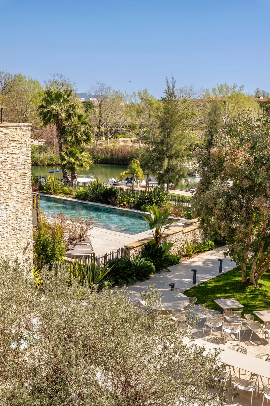 Vue d'un jardin avec piscine, tables et chaises, bordé d'arbres et d'un plan d'eau avec des bateaux