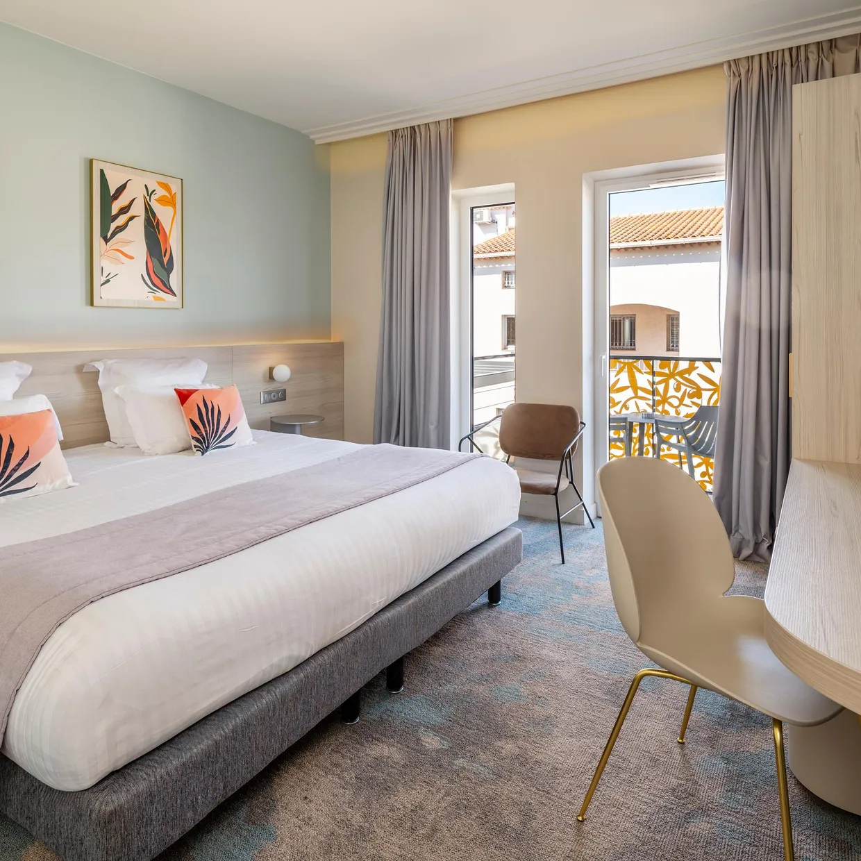 Chambre d'hôtel avec un lit double, une table de chevet, une chaise et une porte-fenêtre donnant sur un balcon avec une vue sur des toits