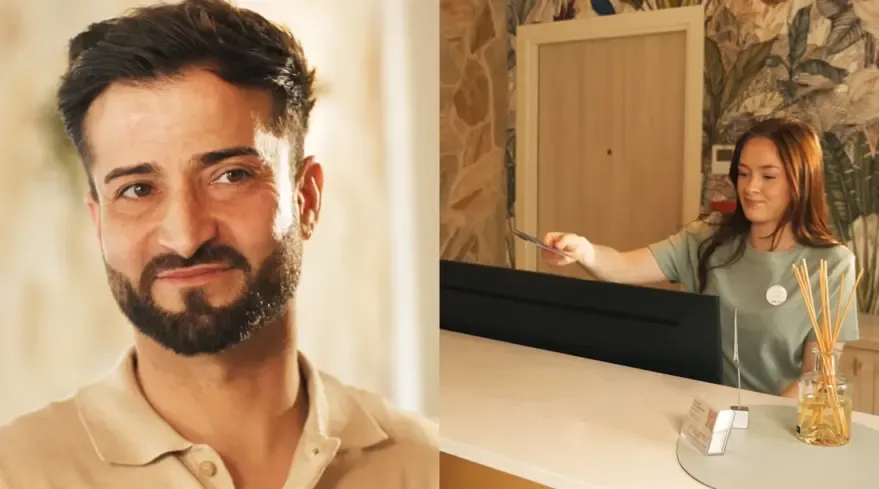 Un homme barbu sourit à gauche, une femme derrière un comptoir tend une carte à droite dans un cadre intérieur avec des plantes en arrière-plan