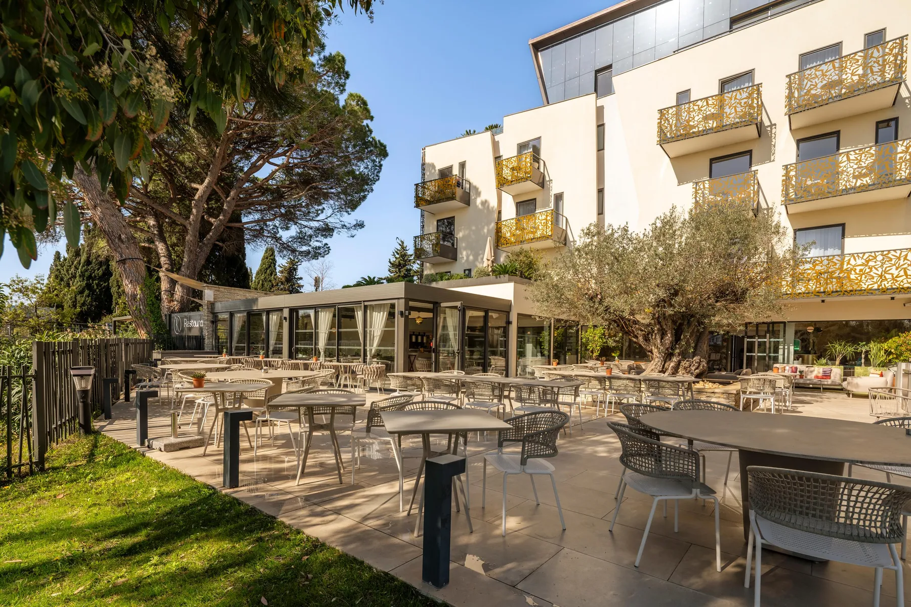 Terrasse extérieure avec tables et chaises, entourée d'arbres et de bâtiments aux balcons ornés de motifs géométriques
