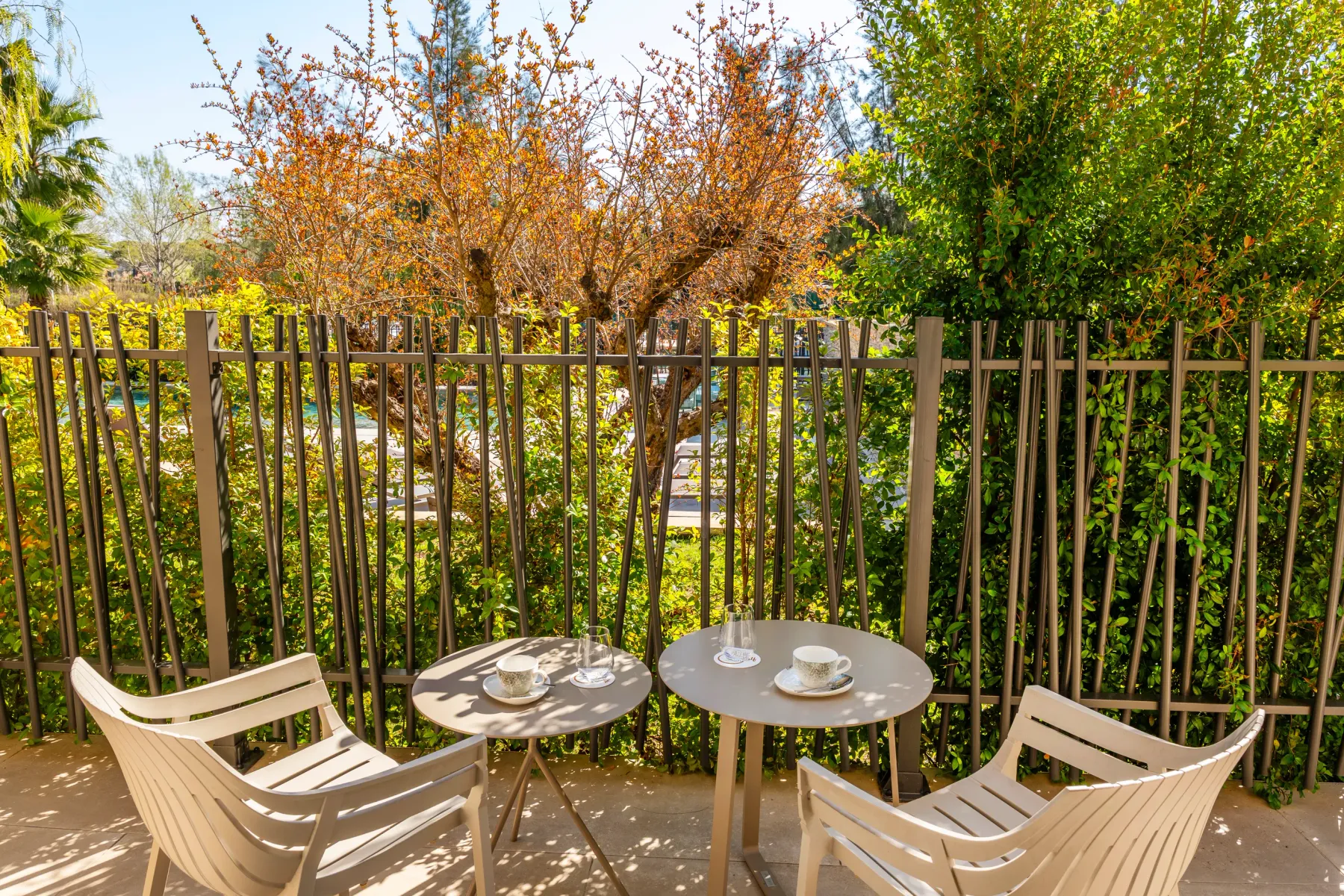 Deux tables rondes avec des tasses à thé et des chaises en bois sur une terrasse entourée d'une végétation dense et d'une clôture en métal