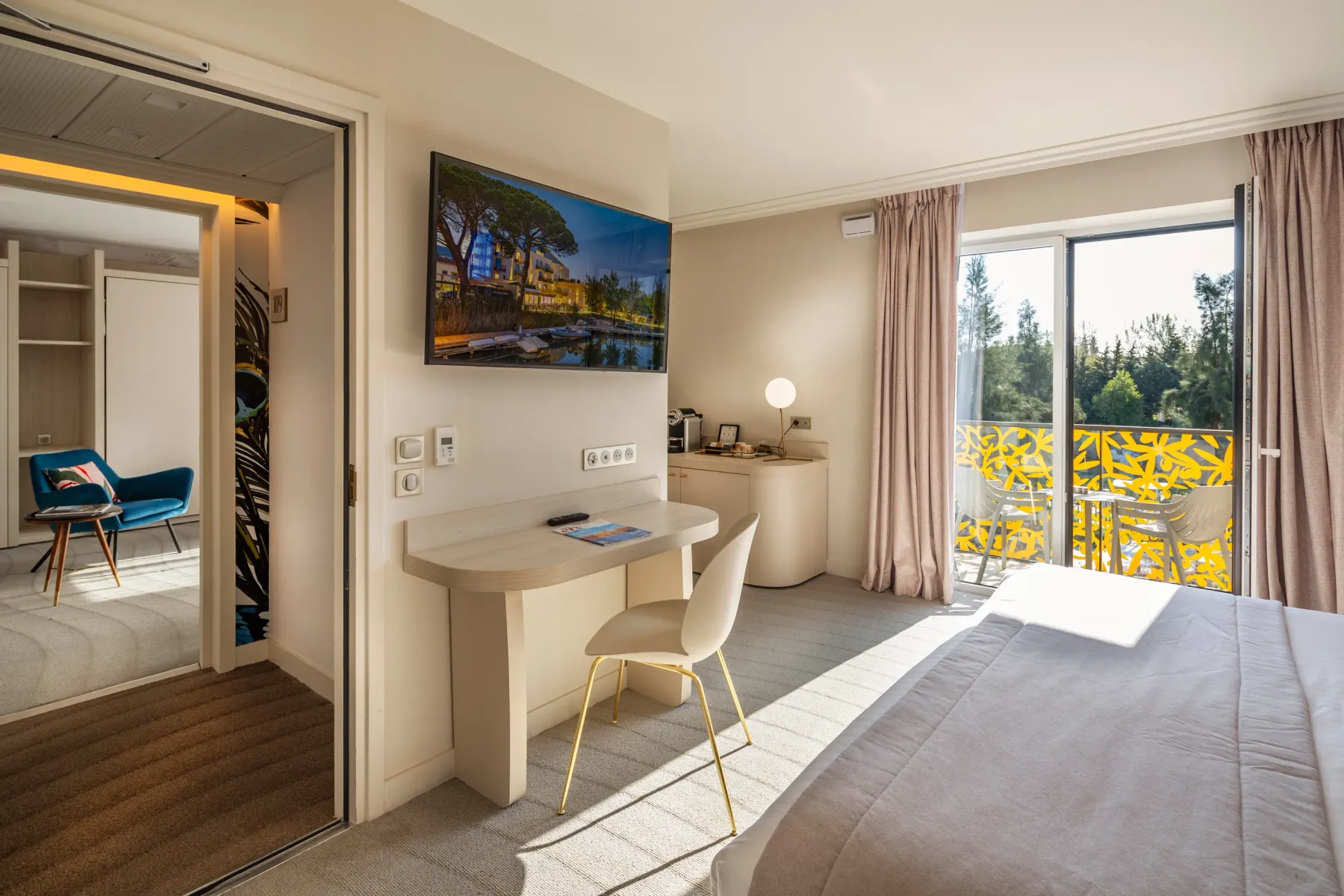 Chambre d'hôtel moderne avec un lit, un bureau, une télévision, un coin salon et une porte-fenêtre donnant sur un balcon avec vue sur la nature