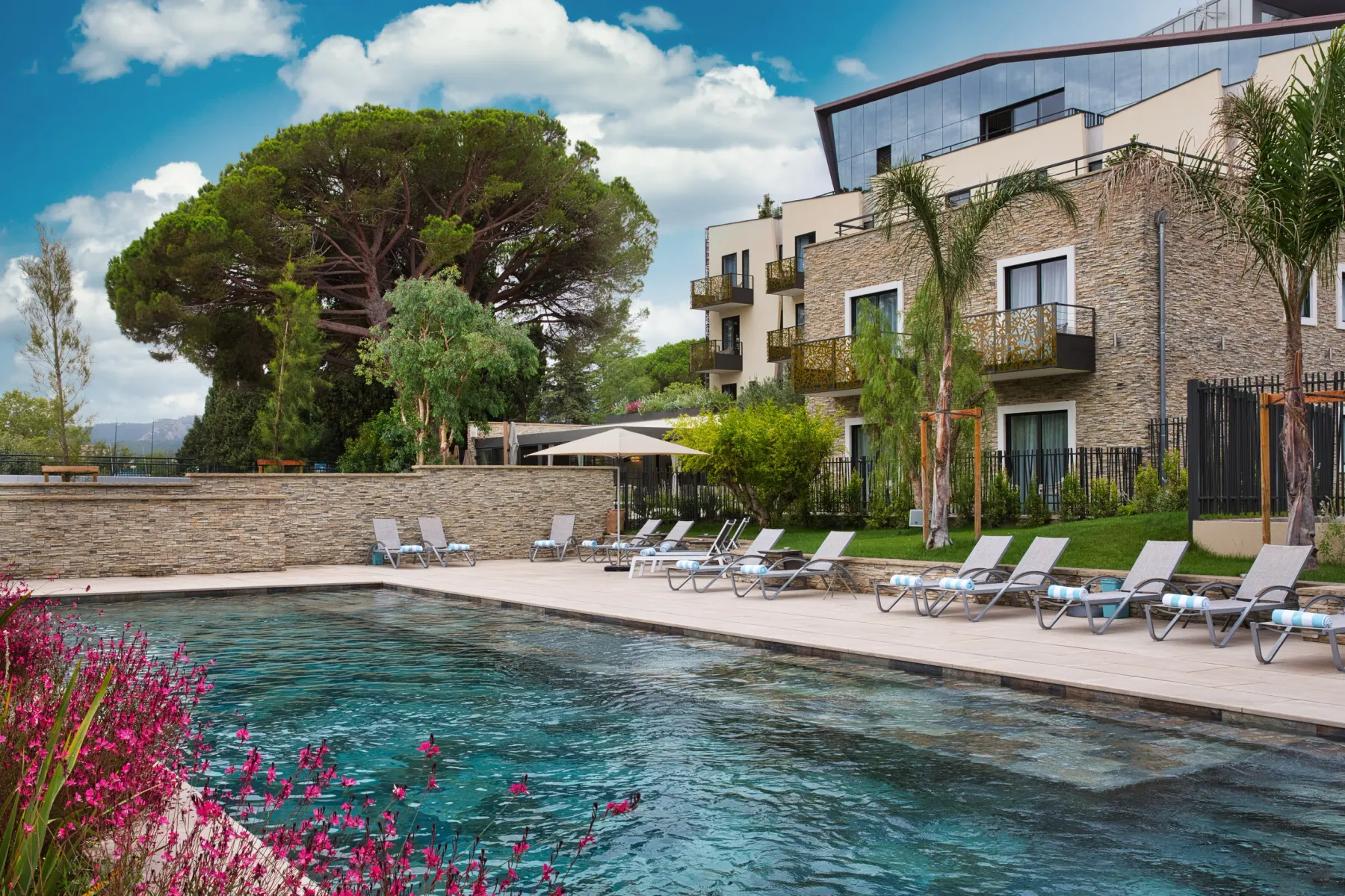 Piscine entourée de chaises longues et d'un bâtiment en pierre avec balcons, sous un ciel partiellement nuageux