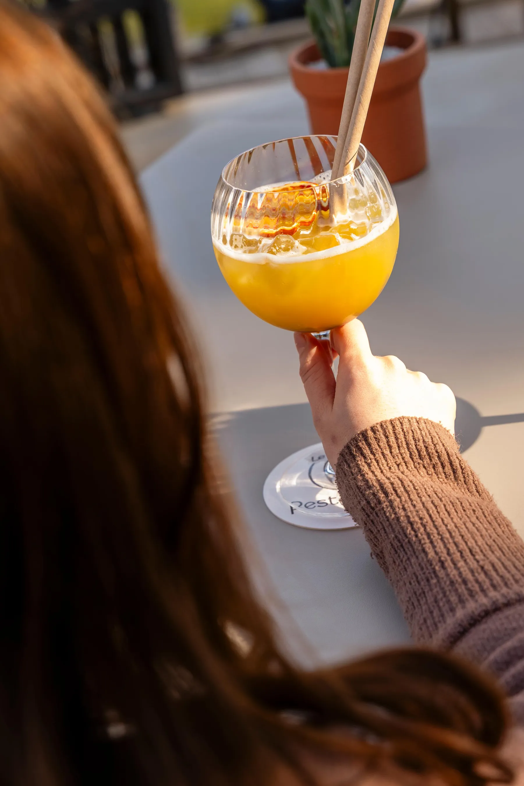 Une main tenant un verre contenant une boisson jaune avec des glaçons et une paille, posée sur une table en extérieur avec des pots de fleurs en arrière-plan
