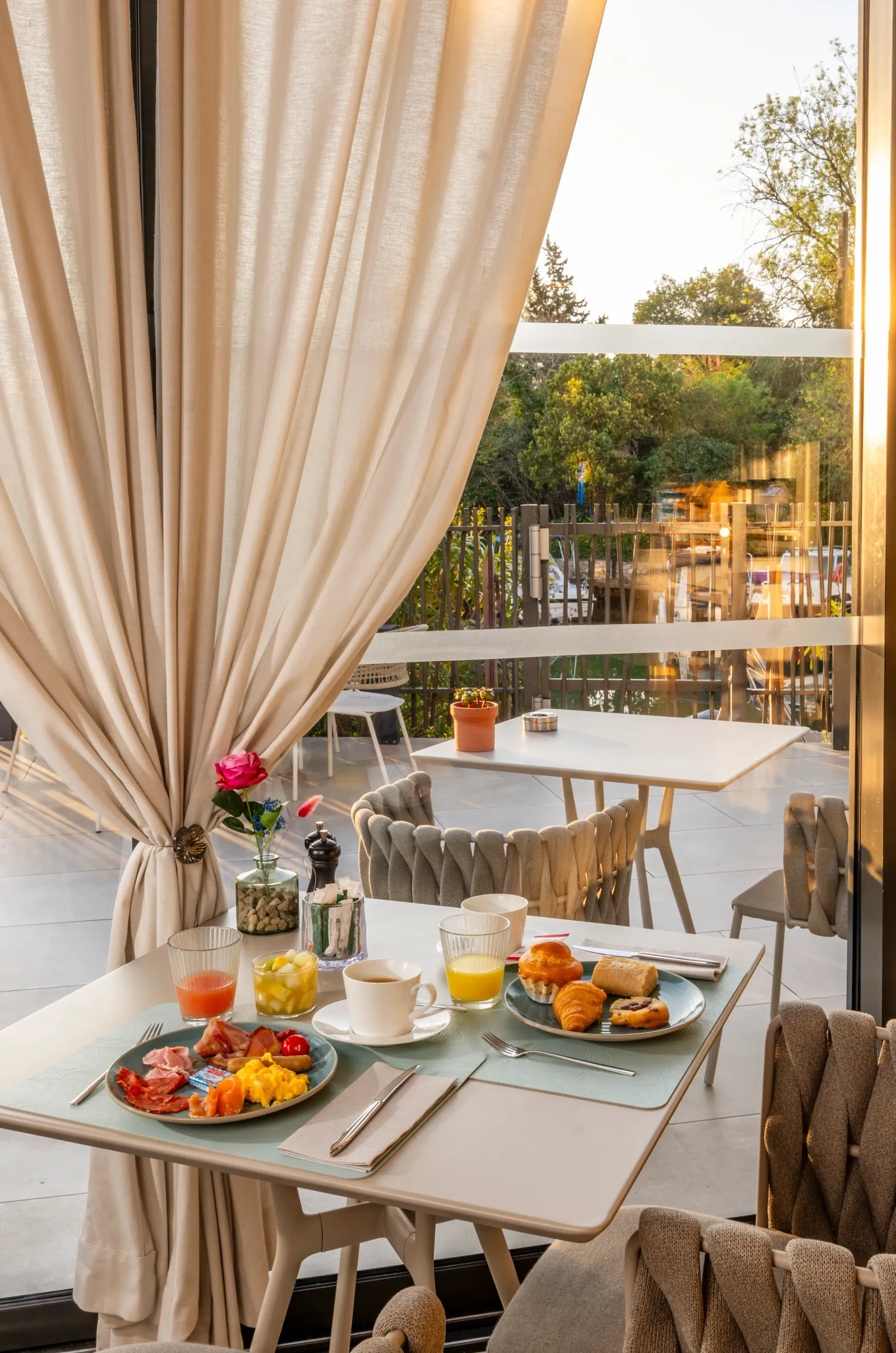 Table dressée pour le petit-déjeuner avec des plats de charcuterie, des croissants, des fruits, des boissons et une rose dans un vase, vue depuis l'intérieur d'un restaurant avec des rideaux et une terrasse extérieure visible à travers une baie vitrée