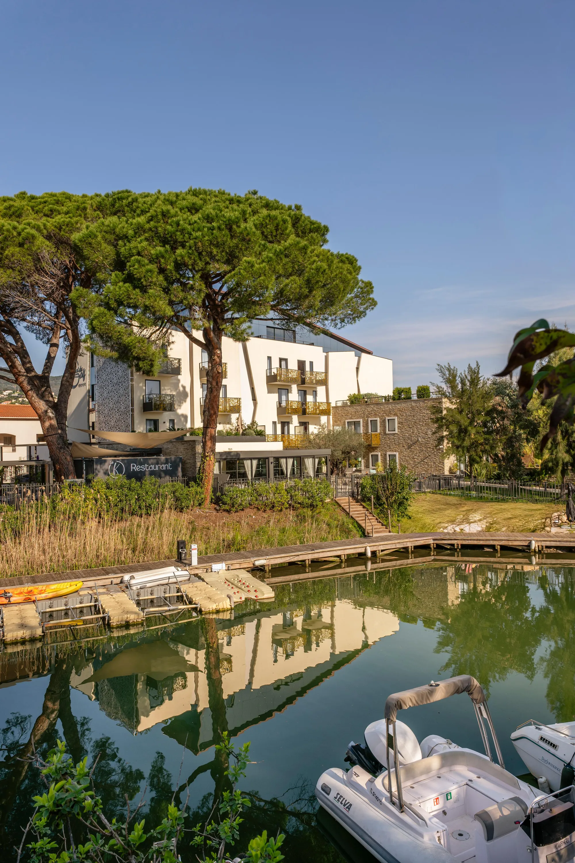 Un canal avec des bateaux amarrés, reflétant un bâtiment blanc et des arbres, entouré de végétation et de structures en bois