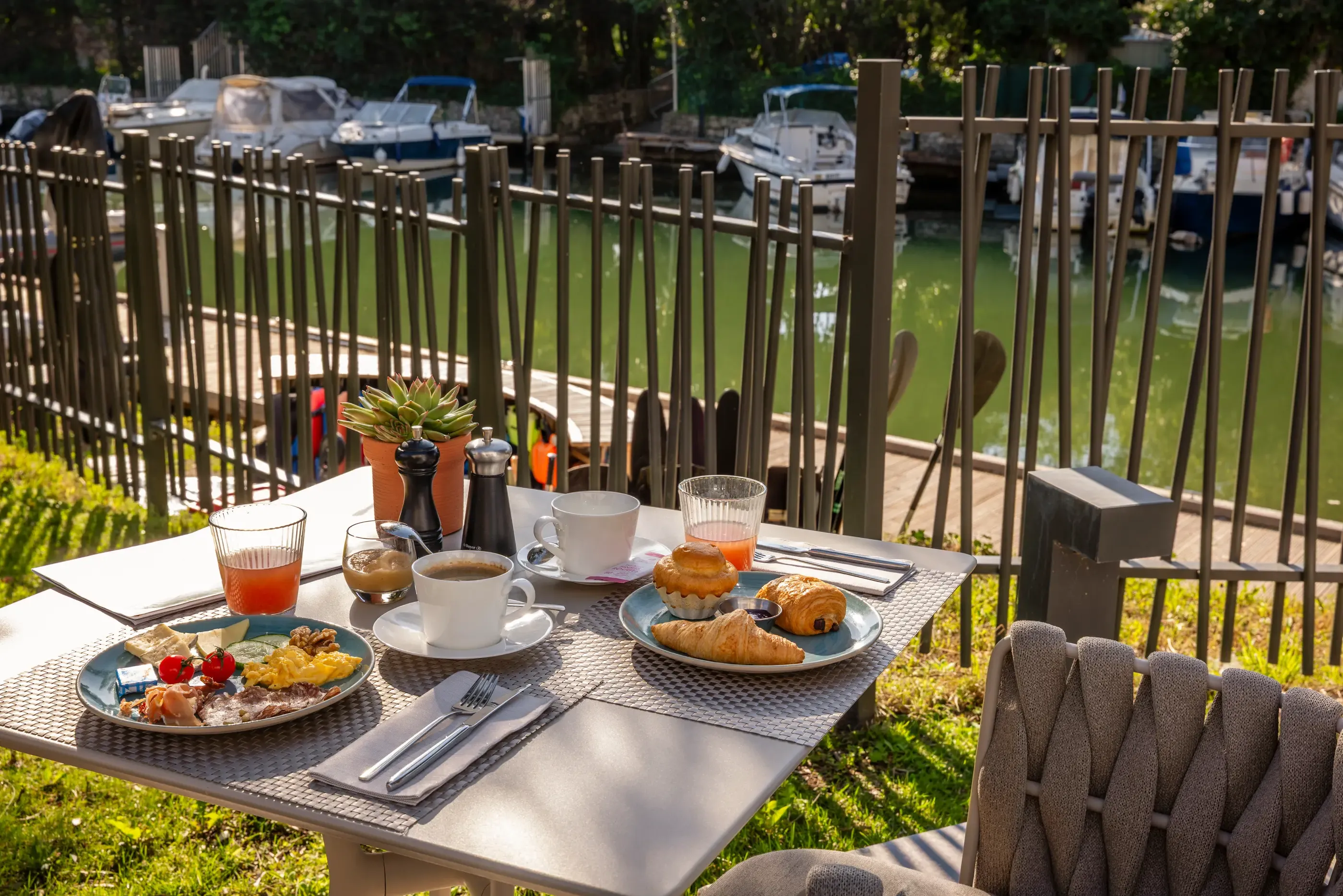 Table en extérieur avec petit-déjeuner composé de croissants, pain, confiture, fruits et boissons, vue sur un canal avec des bateaux