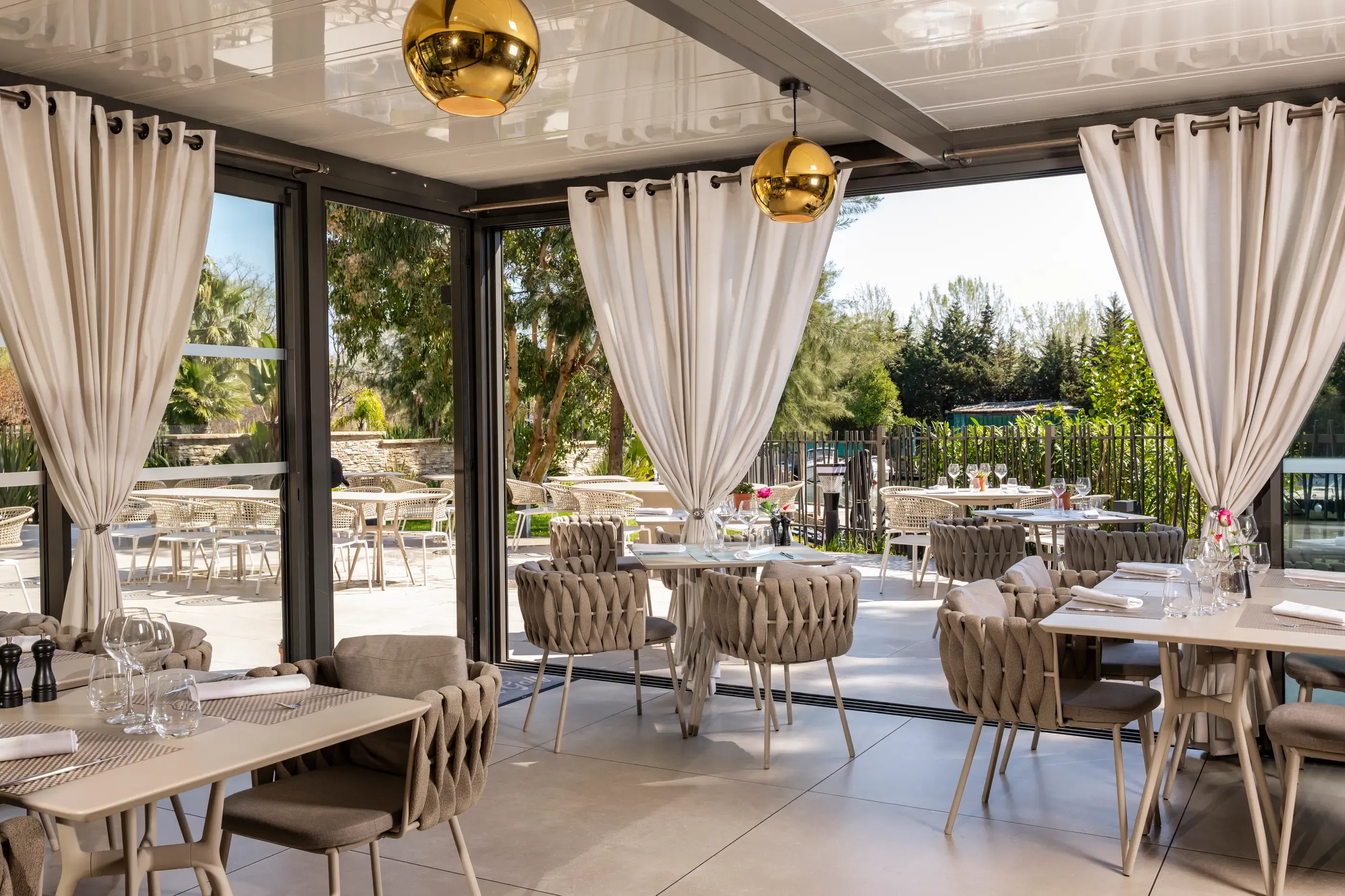Salle de restaurant avec tables et chaises disposées, grandes baies vitrées ouvertes sur une terrasse extérieure, décorée de plantes et de luminaires sphériques