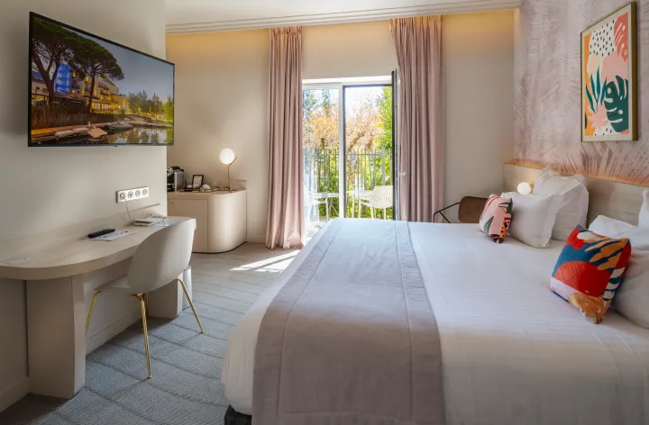 Chambre d'hôtel avec un lit, un bureau, une télévision, une porte-fenêtre donnant sur un balcon et des œuvres d'art aux murs