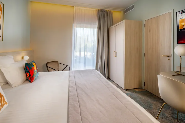 Chambre d'hôtel avec un lit double, une fenêtre avec rideaux, un fauteuil, un bureau et une armoire